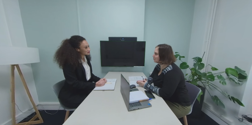 Deux femmes se font face lors d'un recrutement. La candidate est à gauche et parle à la recruteuse, qui est à droite. Elles sont séparées par une table, et il y a une télé au dessus de la table. C'est une capture d'écran d'une des vidéos pour se préparer à un entretien d'embauche.