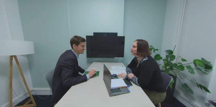 Un homme et une femme se font face lors d'un recrutement. Le candidat est à gauche et parle à la recruteuse, qui est à droite. Ils sont séparés par une table, et il y a une télé au dessus de la table. C'est une capture d'écran d'une des vidéos pour se préparer à un entretien d'embauche.